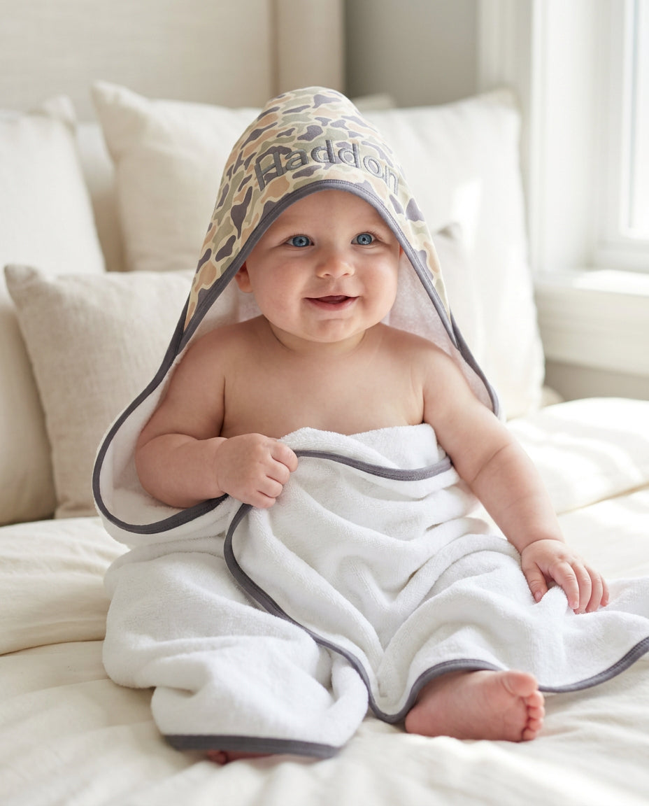 Smiling baby wearing an embroidered bamboo hooded towel with a camo cutie animal print in tan and brown with gray trim