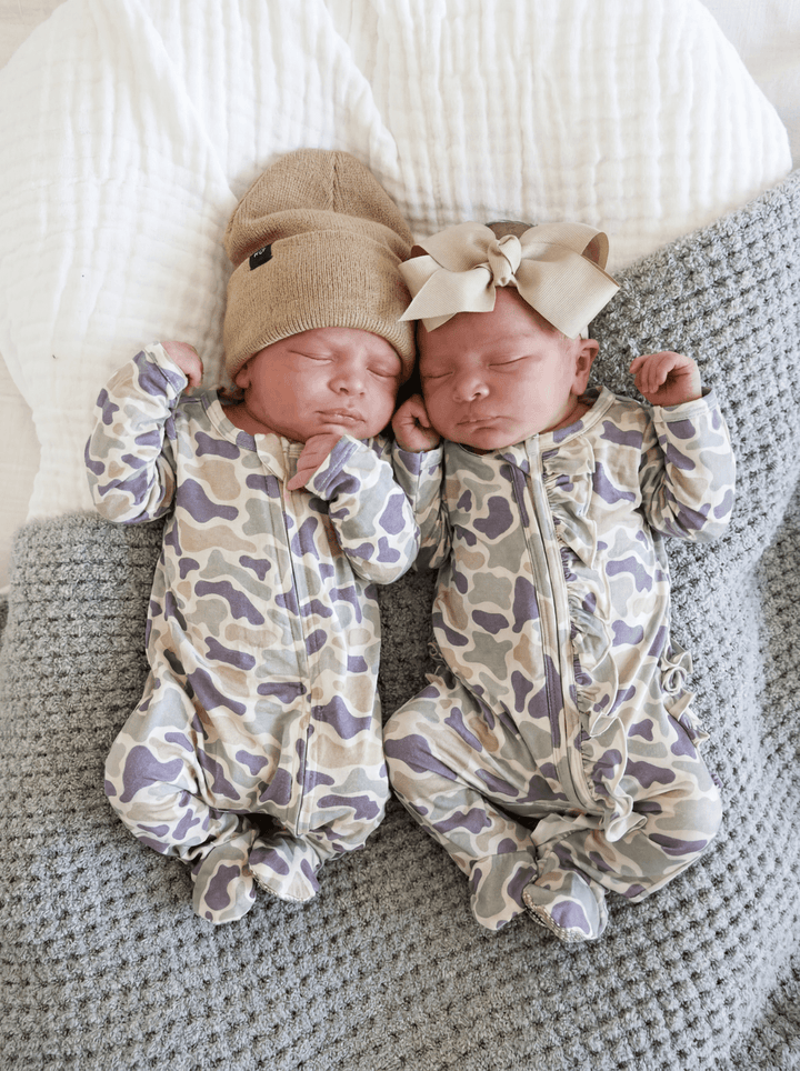 Twin newborn babies lying side by side in matching camo cutie print zipper footies, one with a bear hat and one with a taupe ribbon bow headband