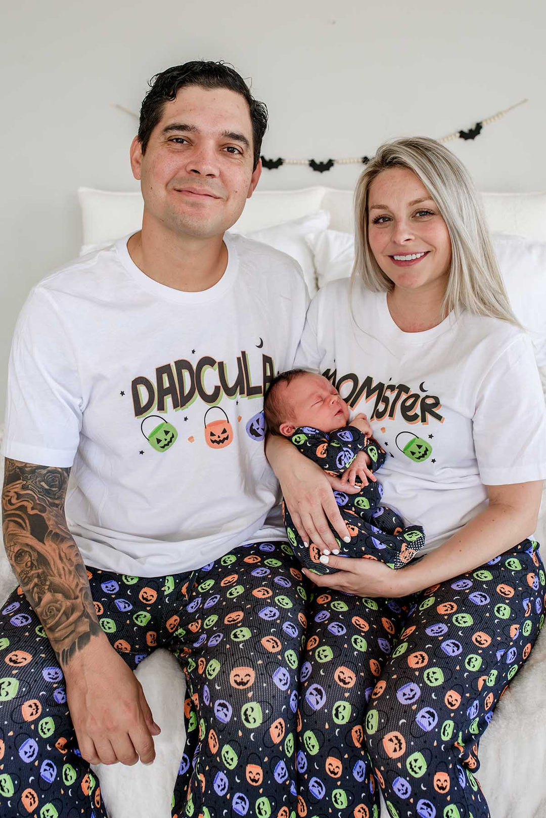Family of three wearing matching Halloween Candy Crawl pajamas: father in 'DADCULA' t-shirt, mother in 'MOMSTER' t-shirt, both in candy-print pajama pants, holding a newborn swaddled in the matching print