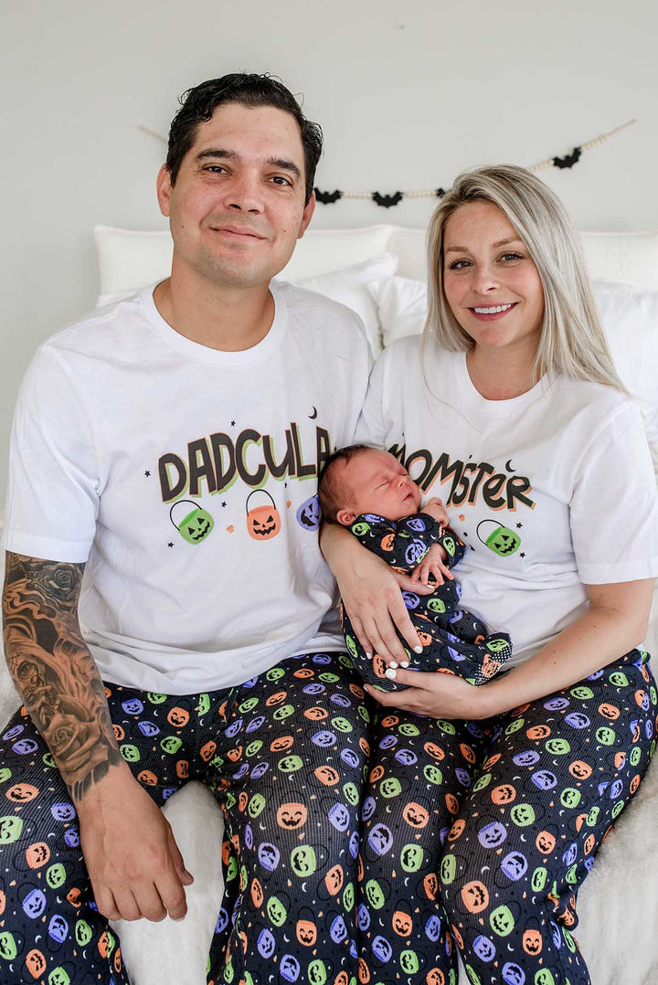 Family of three wearing matching Halloween Candy Crawl pajamas: father in 'DADCULA' t-shirt, mother in 'MOMSTER' t-shirt, both in candy-print pajama pants, holding a newborn swaddled in the matching print