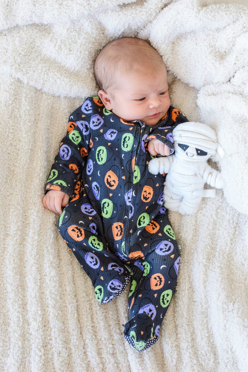 Baby lying on a fluffy blanket in a bamboo waffle zipper footie with colorful jack-o-lantern print, beside a mummy plush toy