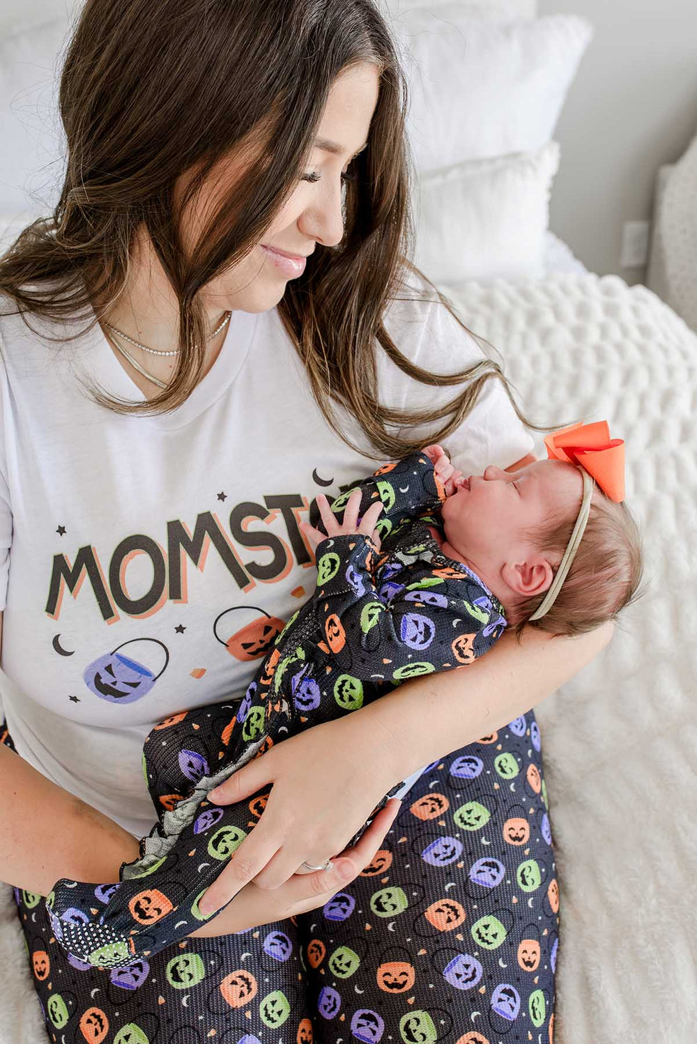 Mother in 'MOMSTER' Halloween t-shirt holding a newborn baby wearing an orange bow headband, swaddled in a candy-print blanket