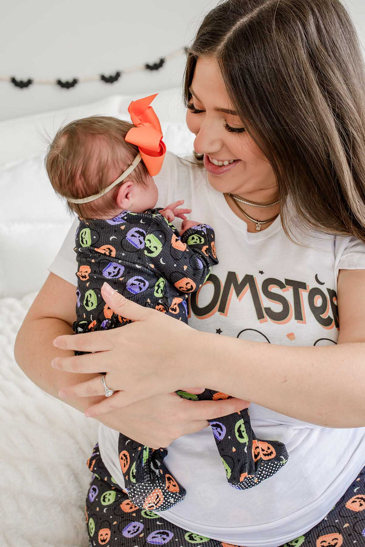 Mother in 'MOMSTER' Halloween t-shirt smiling at newborn baby wearing a large orange bow, swaddled in a candy-print blanket