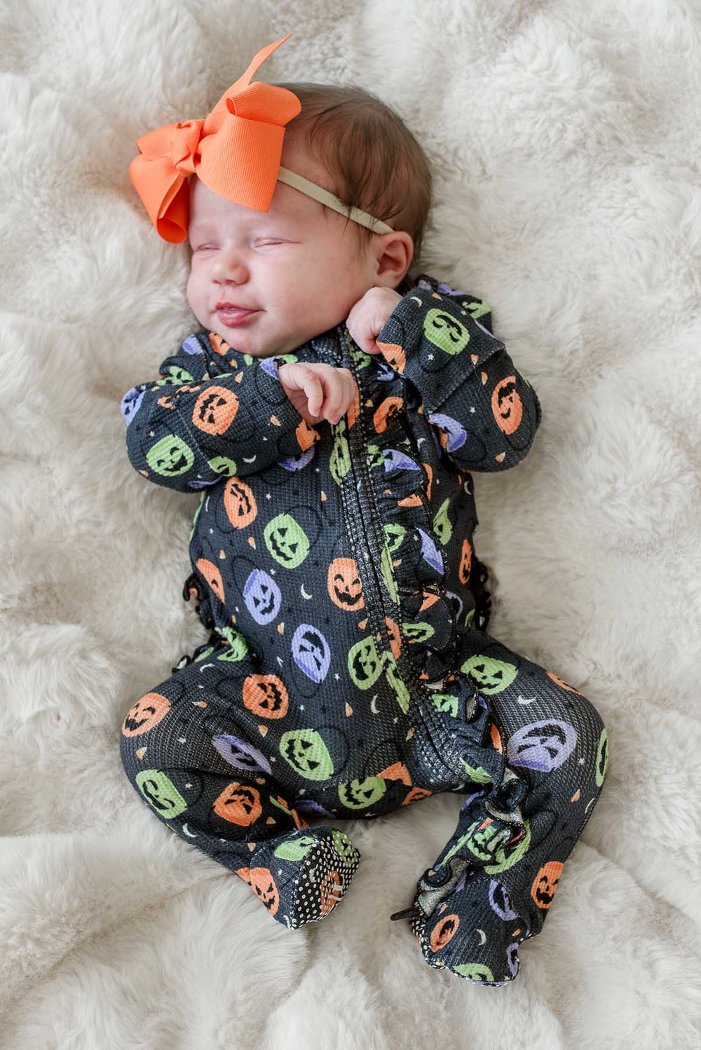 Sleeping newborn with an orange bow wearing a bamboo waffle ruffle footie with colorful jack-o-lantern print, lying on a fluffy blanket