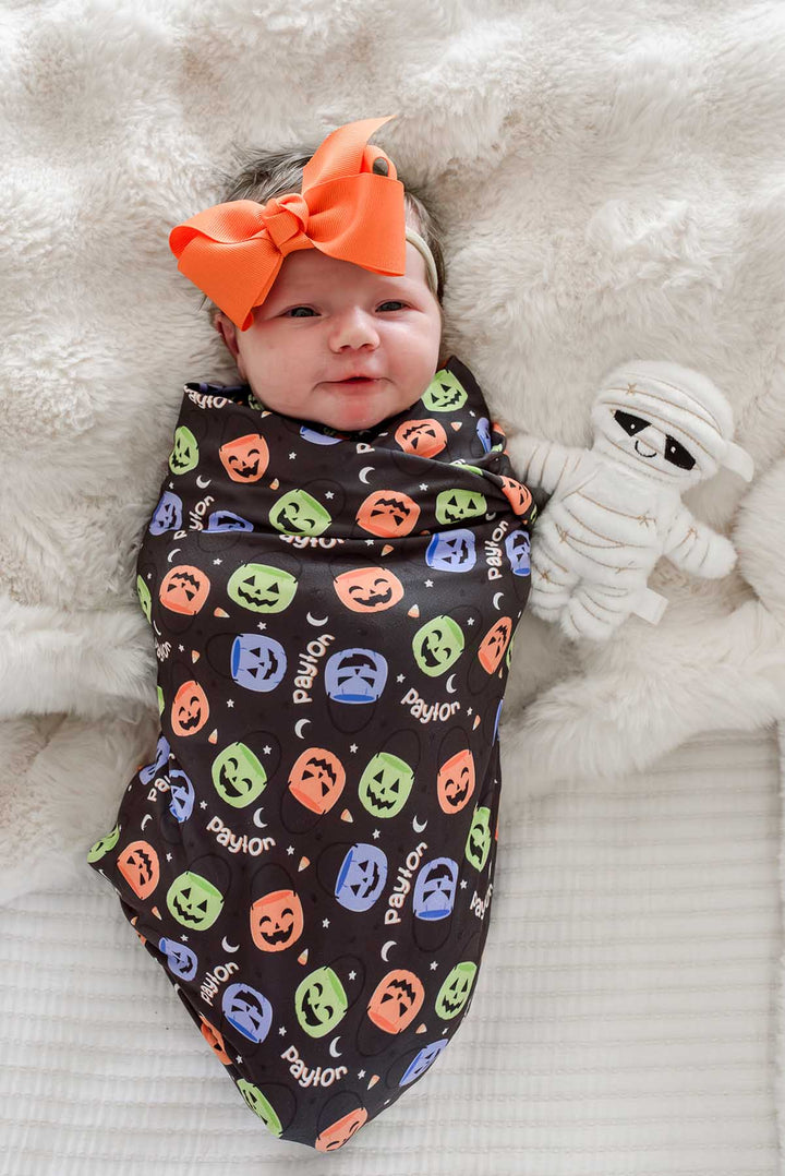 Newborn wearing an orange bow, swaddled in a personalized Halloween blanket with colorful jack-o-lantern print and name text, beside a mummy plush toy