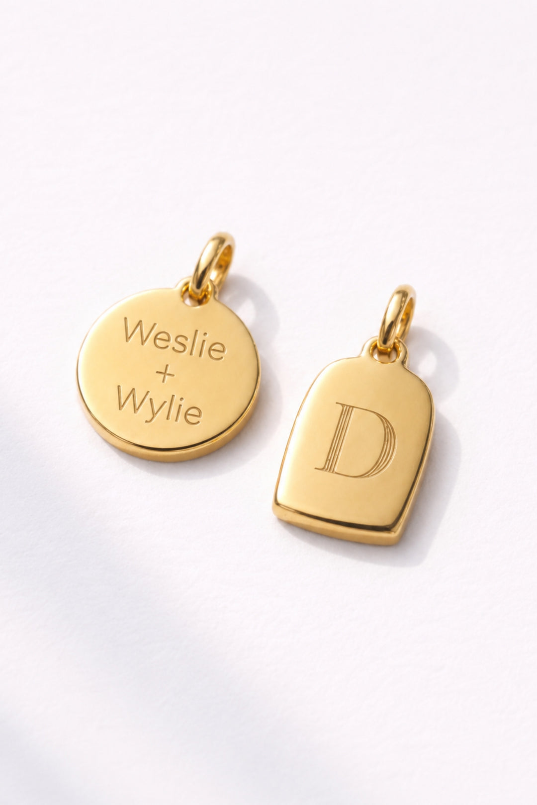 Caden Lane gold charms including engraved "Weslie + Wylie" disc pendant and initial "D" tag charm on white background