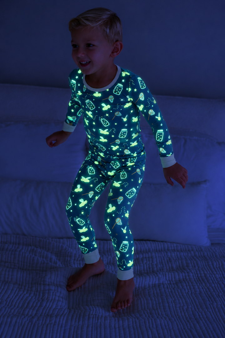 Full-length shot of a boy jumping on a bed, wearing a Caden Lane blue two-piece bamboo pajama set in a Chasing Fireflies print featuring fireflies and mason jars.