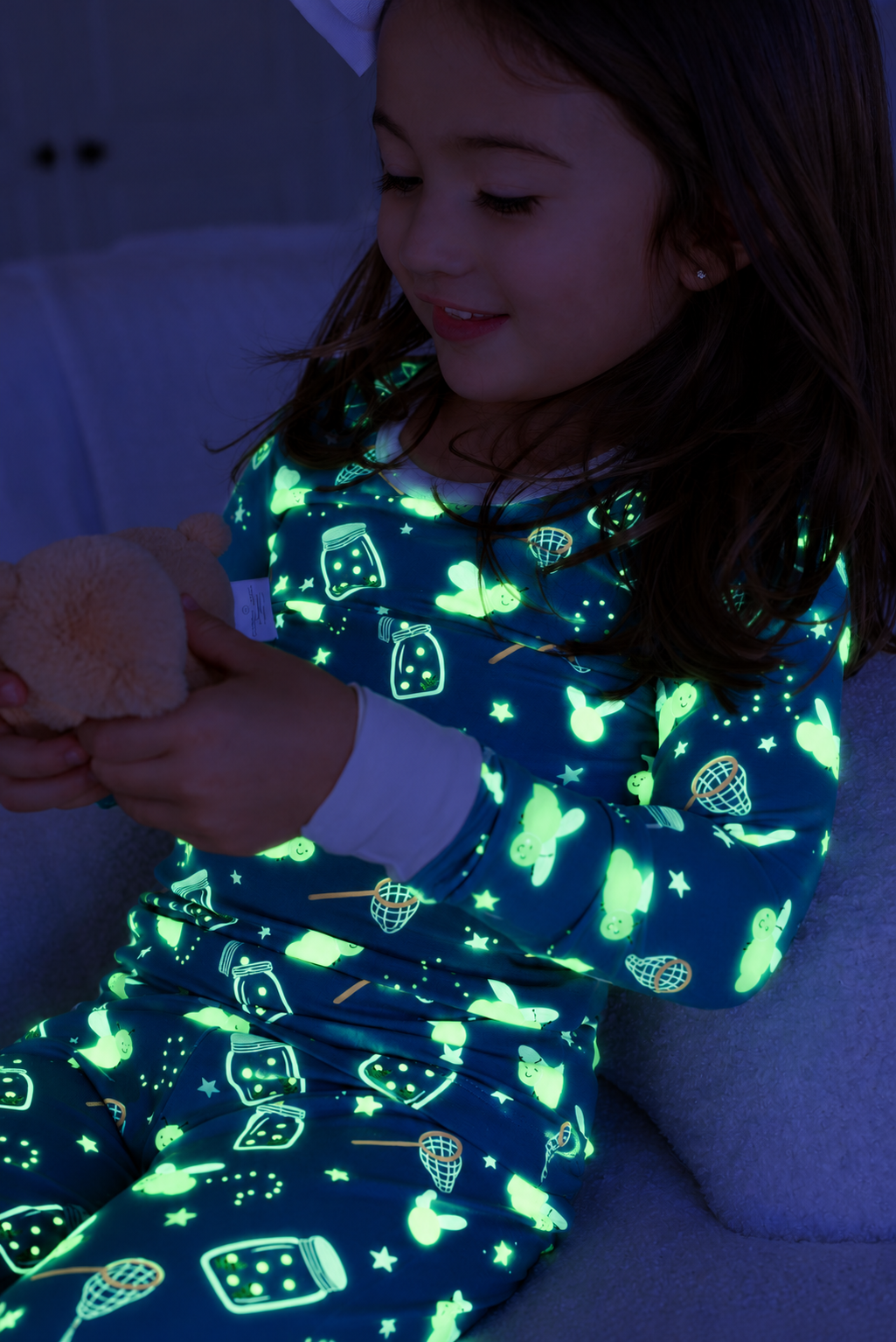 Smiling girl sitting on a couch, wearing a Caden Lane blue two-piece bamboo pajama set in a Chasing Fireflies print featuring fireflies and mason jars, paired with a plush toy.