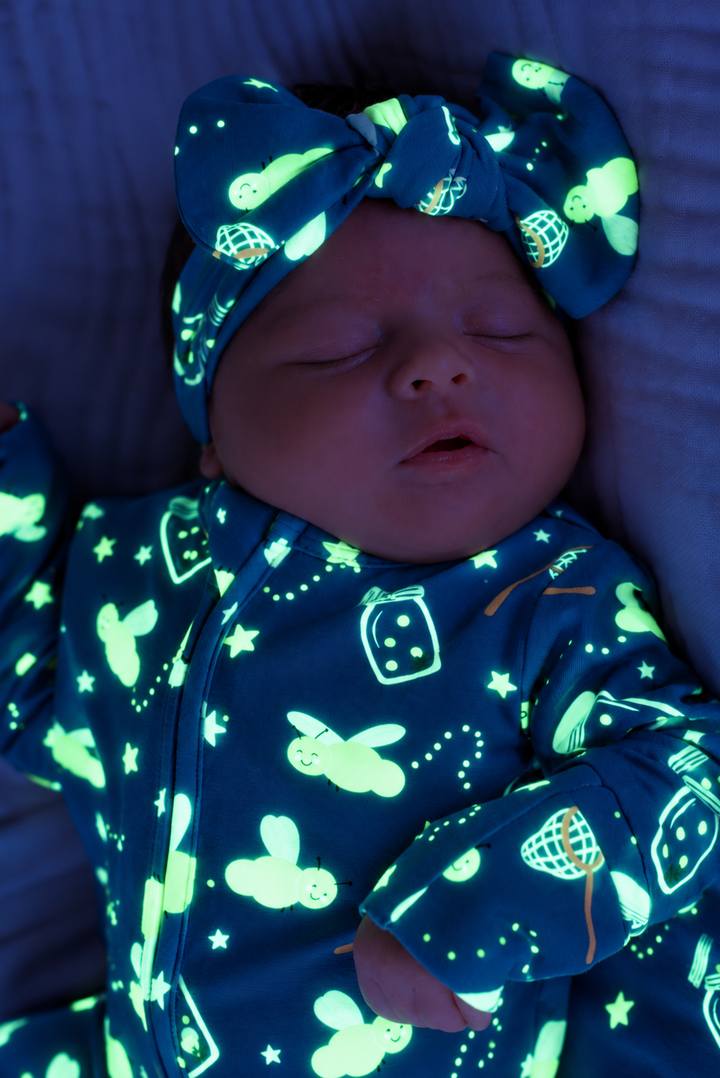 Close-up of a sleeping newborn wearing a Caden Lane blue glow-in-the-dark knit bow headwrap in a Chasing Fireflies print featuring fireflies and mason jars, paired with a matching footie.