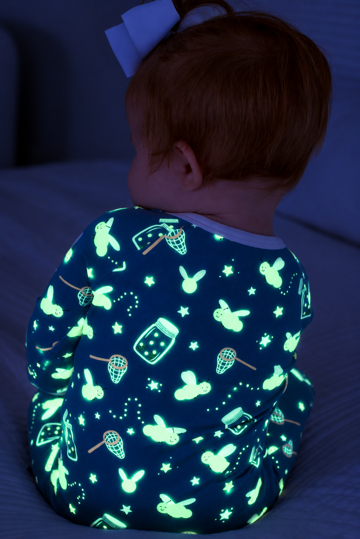 Back view of a seated baby wearing a Caden Lane blue convertible bamboo zip romper in a Chasing Fireflies print featuring fireflies and mason jars, paired with a white bow.