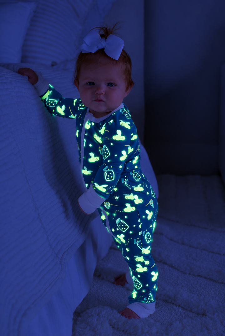 Baby standing, holding onto a bed, wearing a Caden Lane blue convertible bamboo zip romper in a Chasing Fireflies print featuring fireflies and mason jars, paired with a white bow.