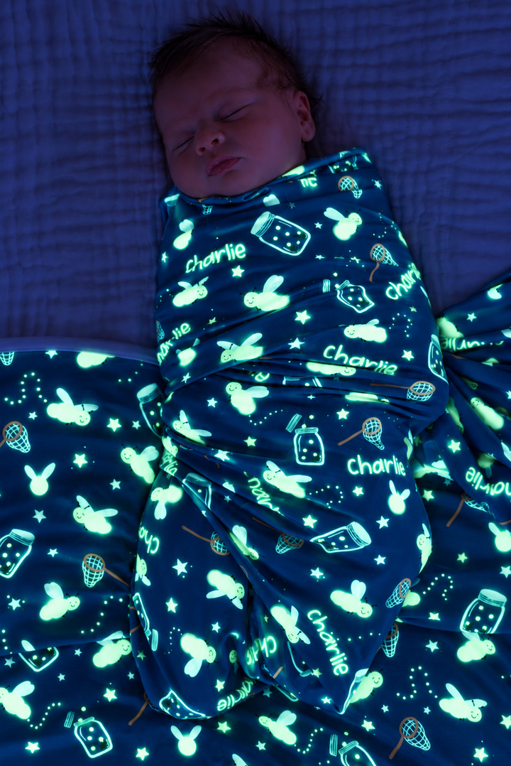 Overhead shot of a sleeping newborn in a dark room, swaddled in a Caden Lane blue personalized swaddle in a Chasing Fireflies print featuring fireflies and mason jars, reading "Charlie."