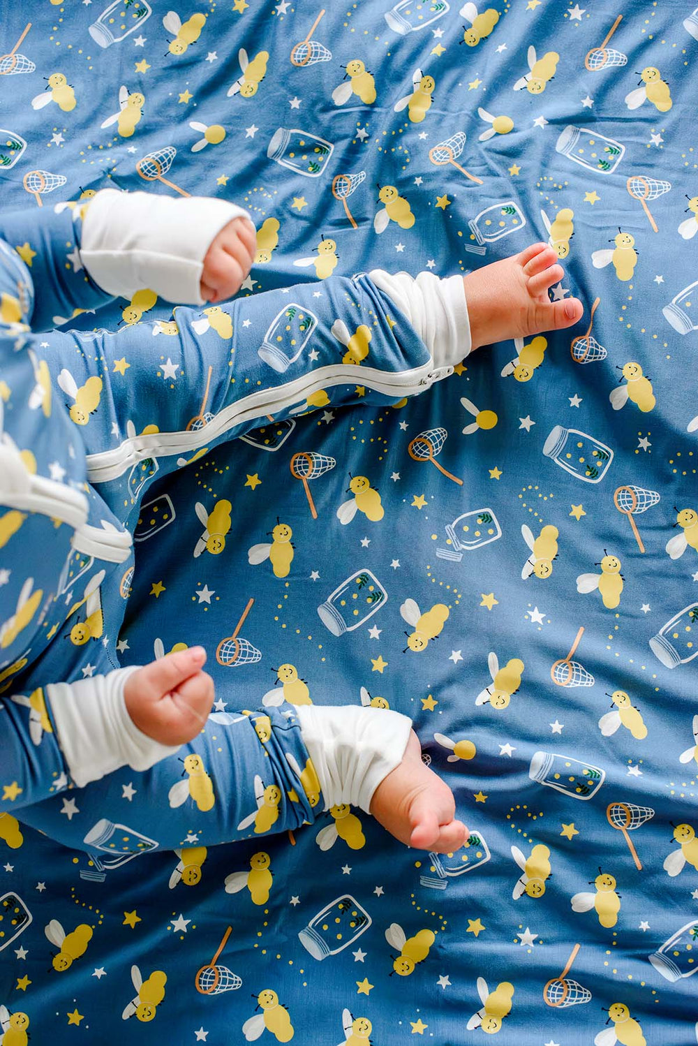 Overhead shot of a baby's feet resting on a Caden Lane blue double-sided bamboo blanket in a Chasing Fireflies print featuring fireflies and mason jars, paired with a matching romper.