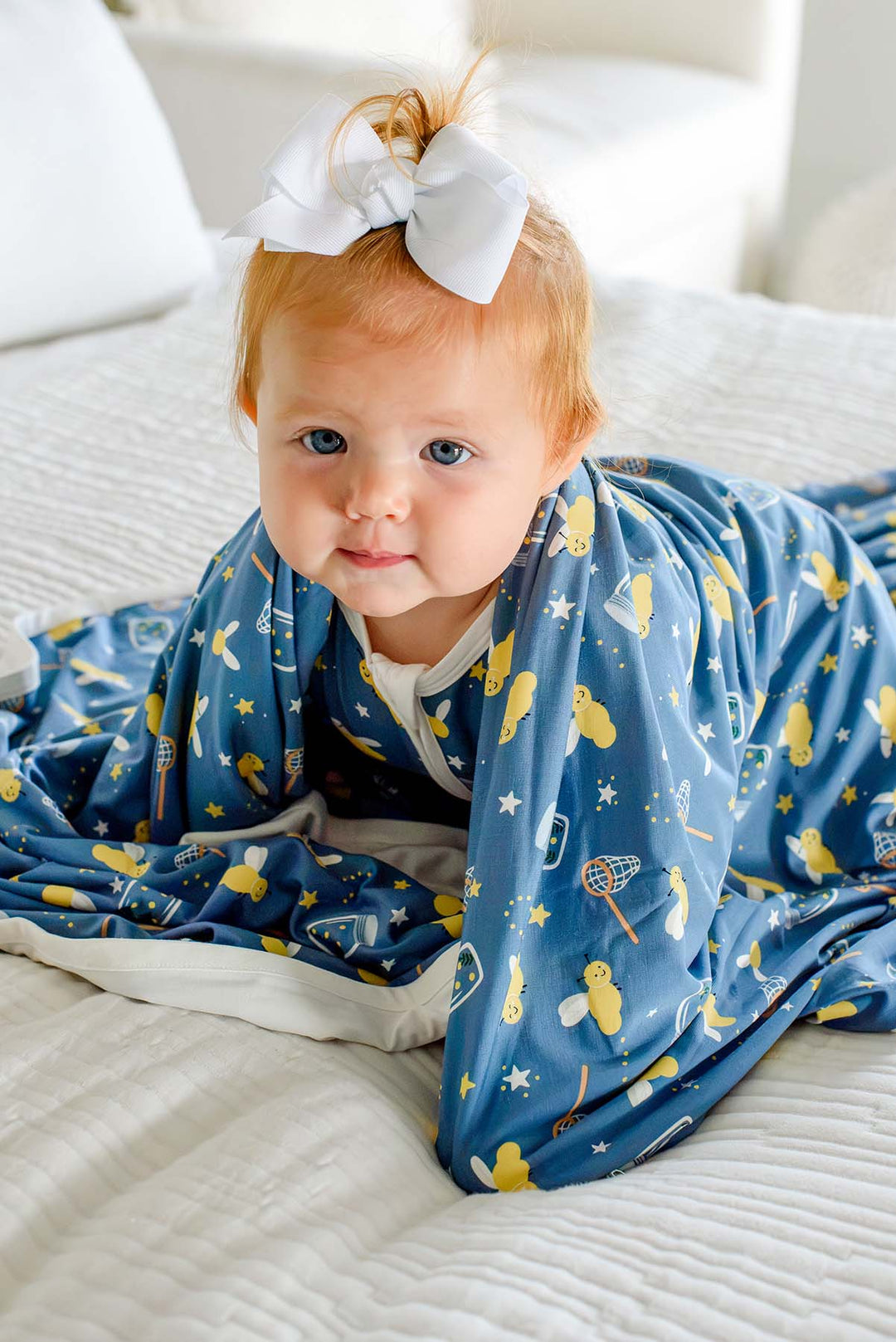 Smiling baby girl crawling out from under a Caden Lane blue double-sided bamboo blanket in a Chasing Fireflies print featuring fireflies and mason jars, paired with a white hair bow.