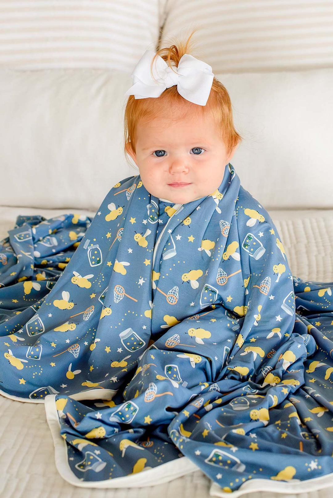 Red-haired baby girl sitting wrapped in a Caden Lane blue double-sided bamboo blanket in a Chasing Fireflies print featuring fireflies and mason jars, paired with a white hair bow.
