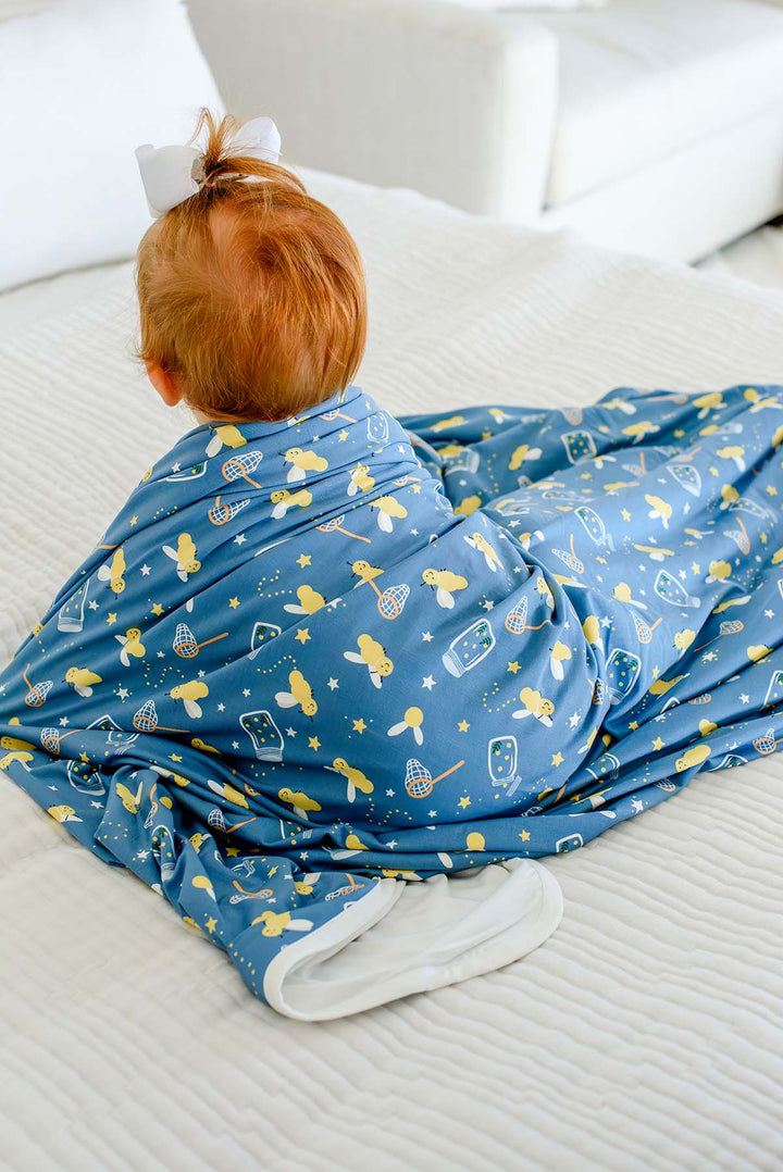 Back view of a red-haired baby draped in a Caden Lane blue double-sided bamboo blanket in a Chasing Fireflies print featuring fireflies and mason jars, showing the cream reverse side.