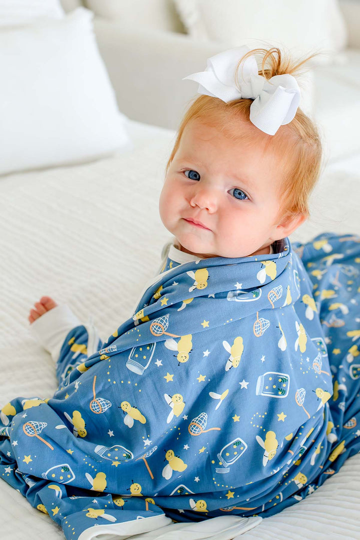 Baby girl glancing over her shoulder, wrapped in a Caden Lane blue double-sided bamboo blanket in a Chasing Fireflies print featuring fireflies and mason jars, paired with a white bow.