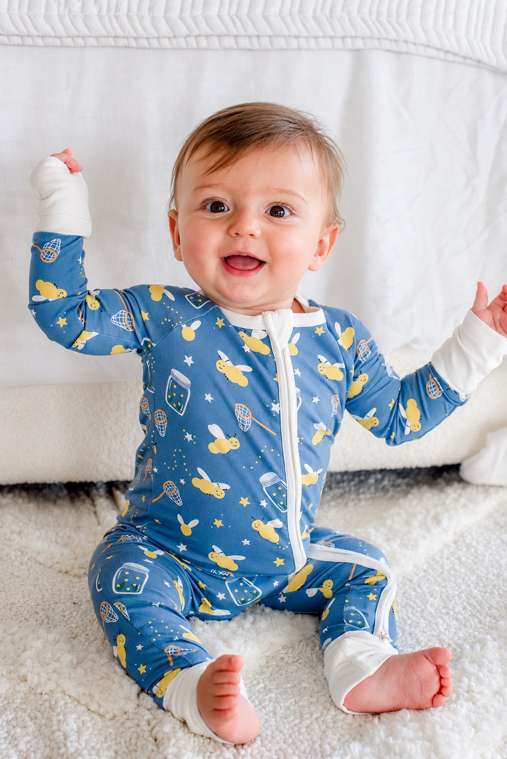 Baby boy sitting on a fluffy rug sticking out his tongue, wearing a Caden Lane blue convertible bamboo zip romper in a Chasing Fireflies print featuring fireflies and mason jars.