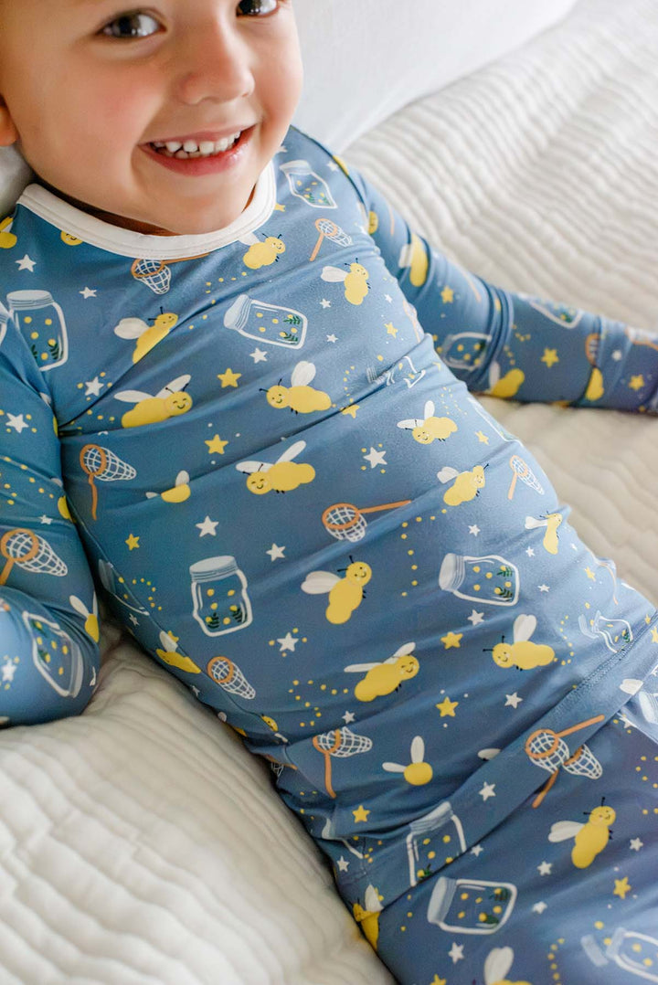 Close-up of a smiling child lounging on pillows wearing a Caden Lane blue two-piece bamboo pajama set in a Chasing Fireflies print featuring fireflies and mason jars.