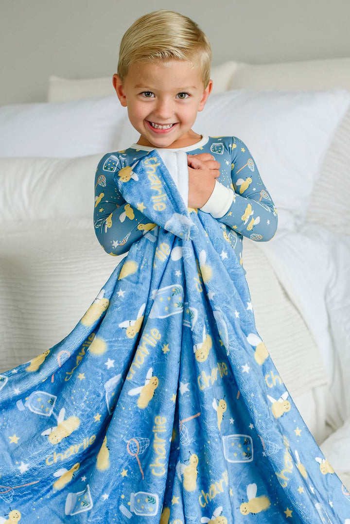 Grinning toddler boy hugging a Caden Lane blue plush personalized blanket to his chest in a Chasing Fireflies print featuring fireflies and mason jars, reading "Charlie," paired with matching pajamas.