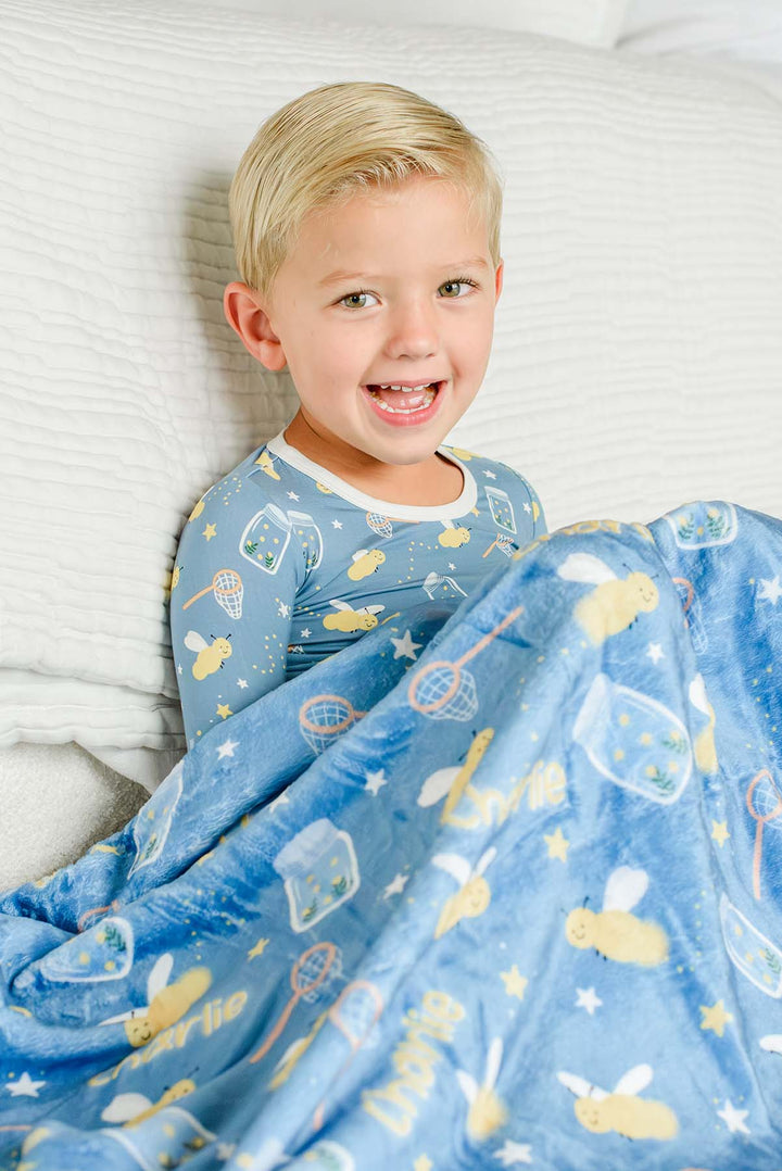 Laughing toddler boy leaning on pillows under a Caden Lane blue plush personalized blanket in a Chasing Fireflies print featuring fireflies and mason jars, paired with matching pajamas.