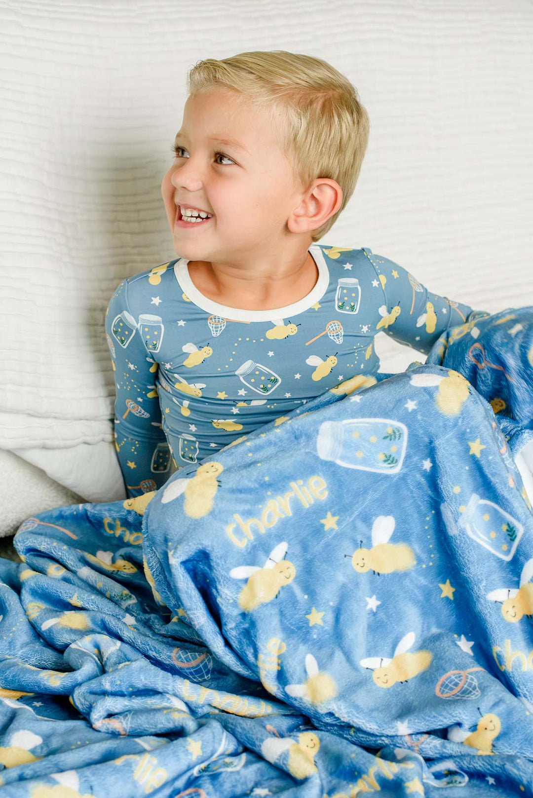Toddler boy glancing sideways under a Caden Lane blue plush personalized blanket in a Chasing Fireflies print featuring fireflies and mason jars, reading "Charlie," paired with matching pajamas.