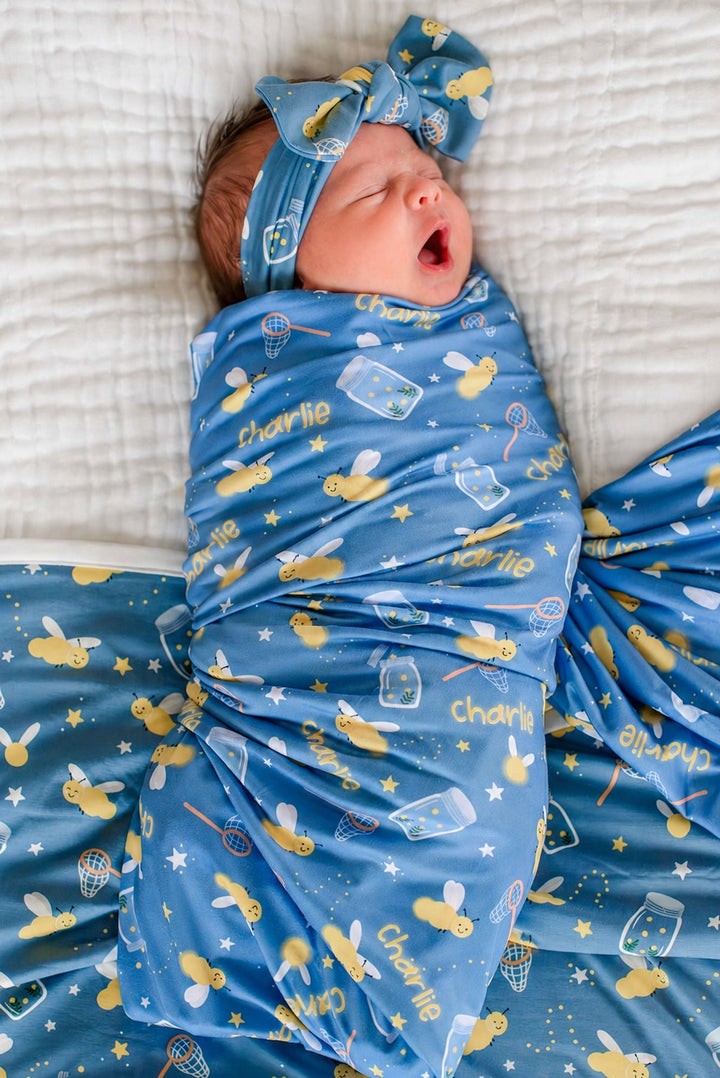 Overhead shot of a swaddled newborn in a Caden Lane blue personalized swaddle in a Chasing Fireflies print featuring fireflies and mason jars, reading "Charlie," paired with a bow headwrap.