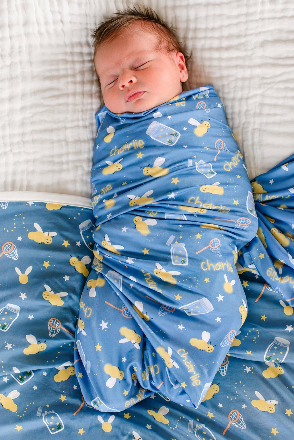 Overhead shot of a wide-eyed newborn bundled in a Caden Lane blue personalized swaddle in a Chasing Fireflies print featuring fireflies and mason jars, reading "Charlie."