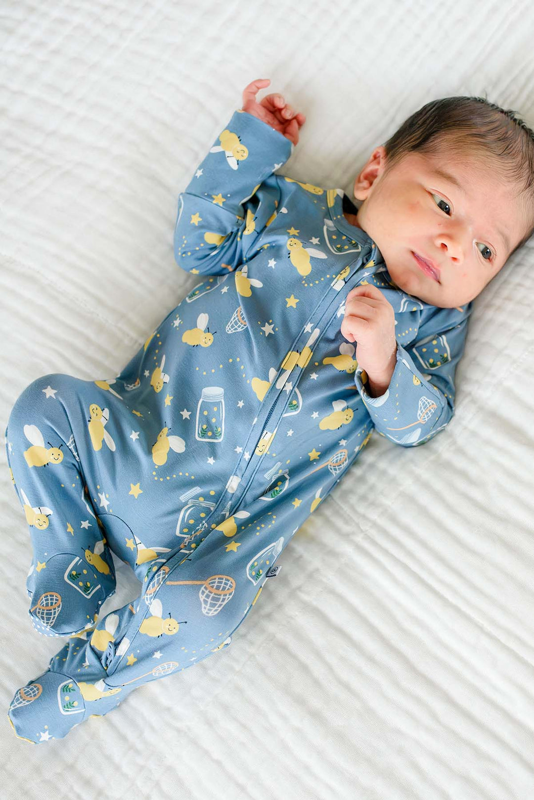 Overhead shot of a newborn lying on a white blanket wearing a Caden Lane blue bamboo zipper footie in a Chasing Fireflies print featuring fireflies and mason jars.