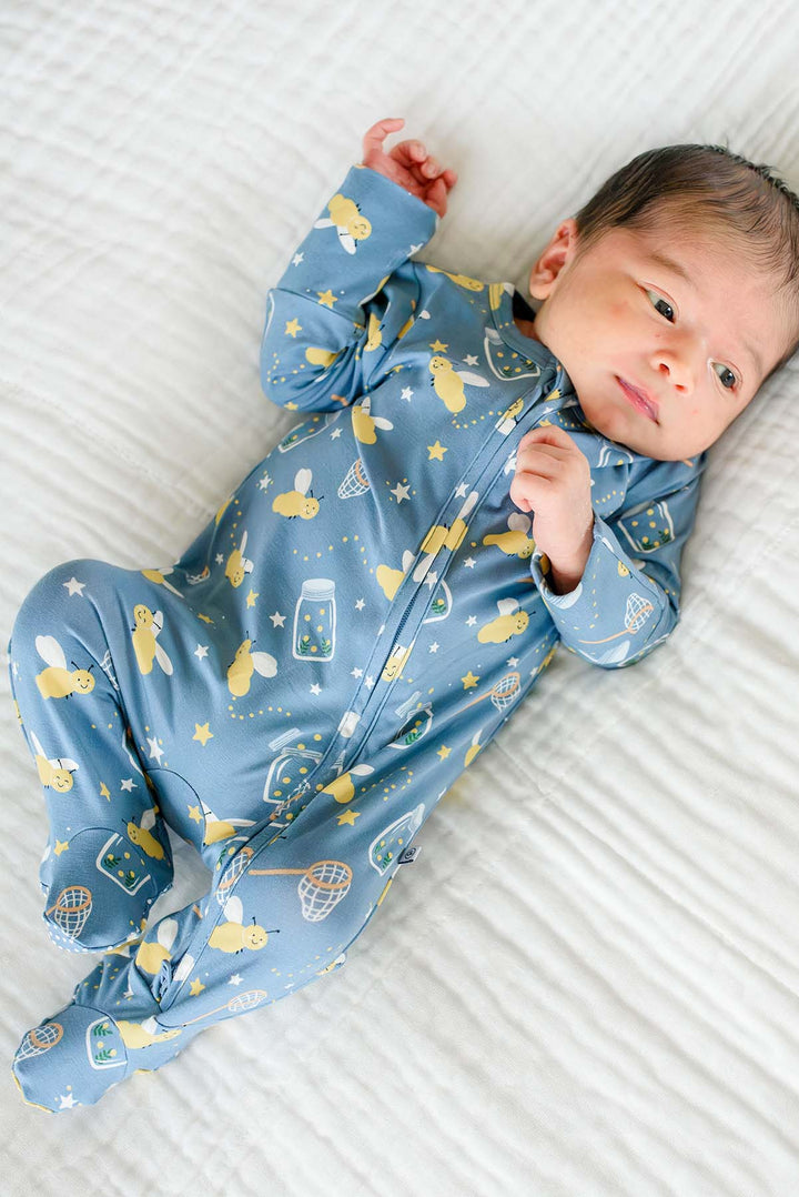 Overhead shot of a newborn lying on a white blanket wearing a Caden Lane blue bamboo zipper footie in a Chasing Fireflies print featuring fireflies and mason jars.