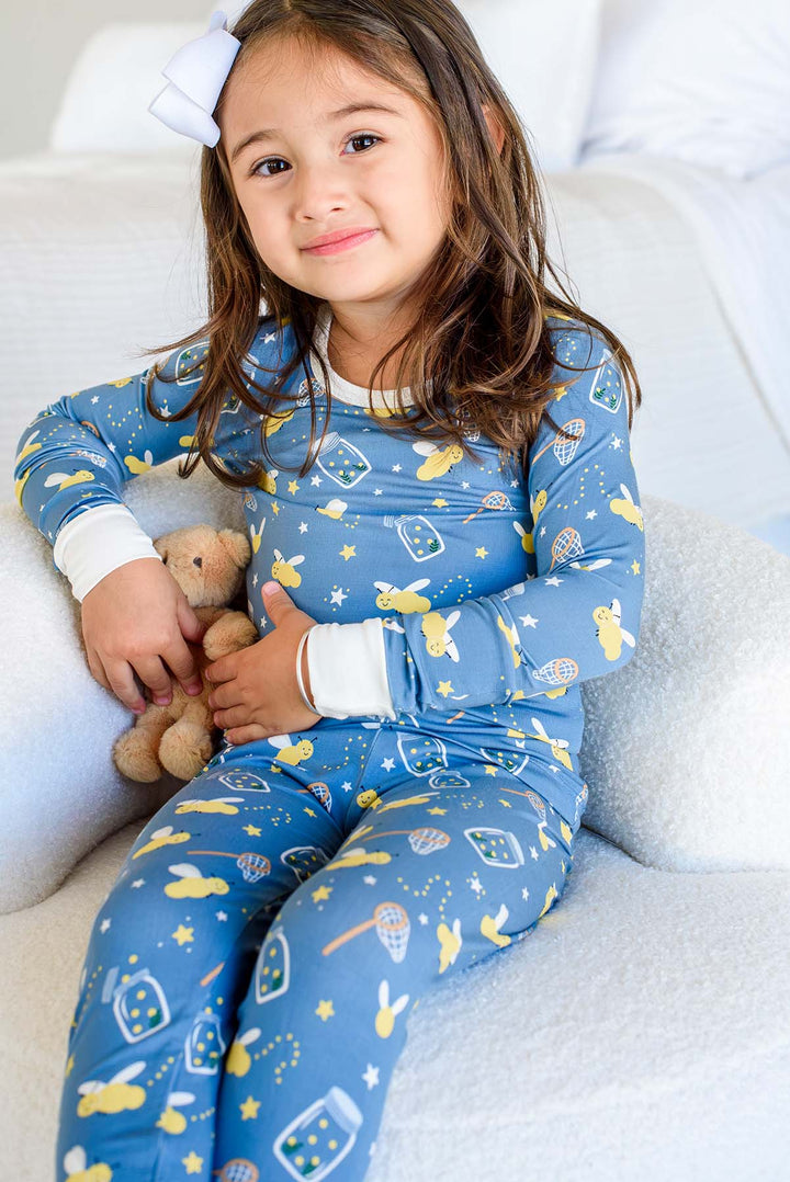 Girl sitting in a bouclé chair wearing a Caden Lane blue two-piece bamboo pajama set in a Chasing Fireflies print featuring fireflies and mason jars, paired with a teddy bear.