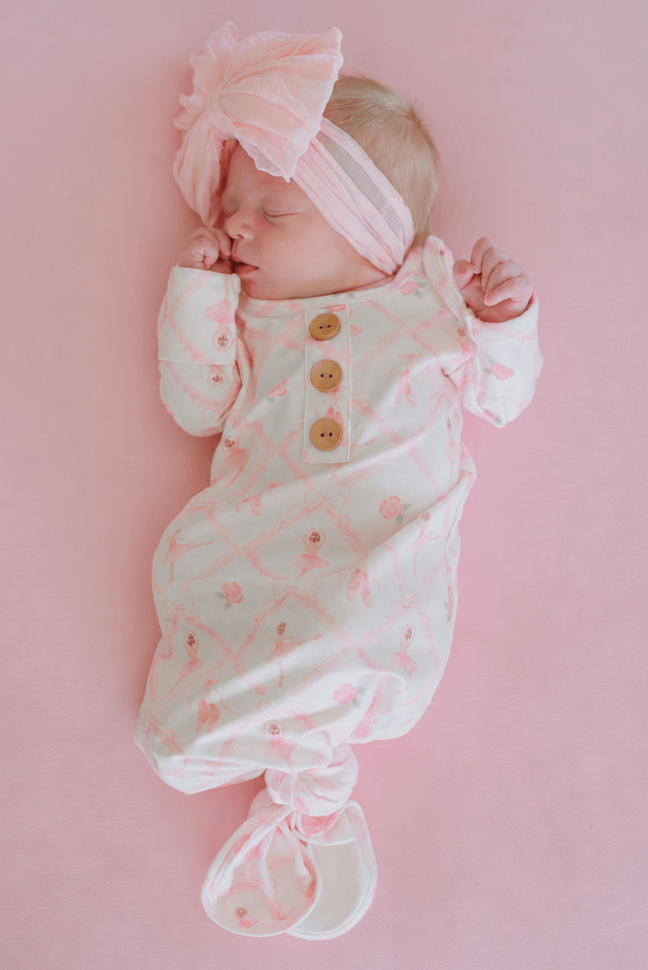 Newborn baby lying on a pink backdrop wearing a Caden Lane ballerina and roses pink print knotted gown with wooden buttons and matching oversized bow headband