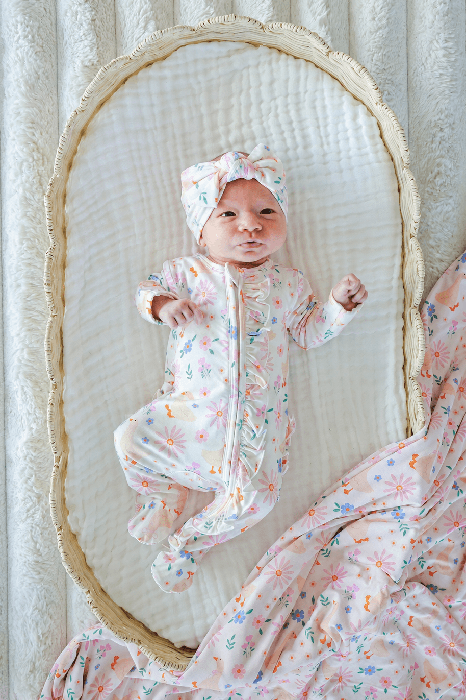 Newborn baby in a bassinet wearing the Lucy Goosy floral print outfit and Knit Large Bow Headwrap, lifestyle overhead photo showing the complete look