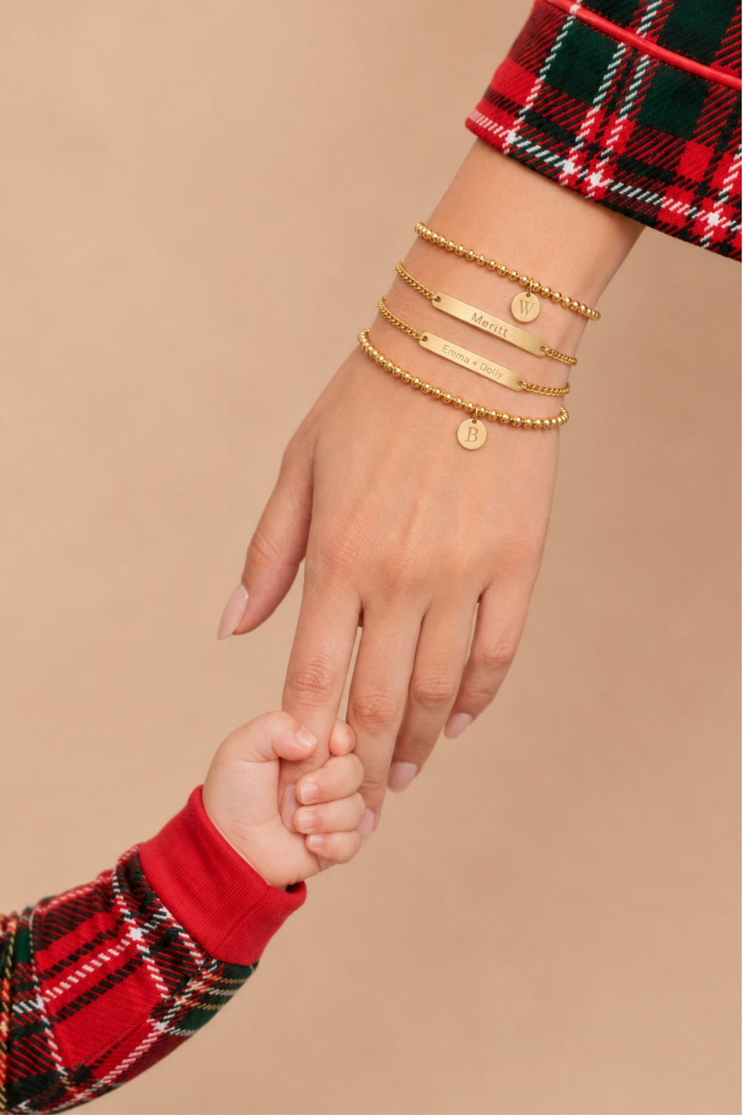 Caden Lane layered gold bracelets with "Meritt" and "Emma + Dolly" engraved bars and initial "W" and "B" discs, styled with plaid holiday pajamas, mother and child holding hands