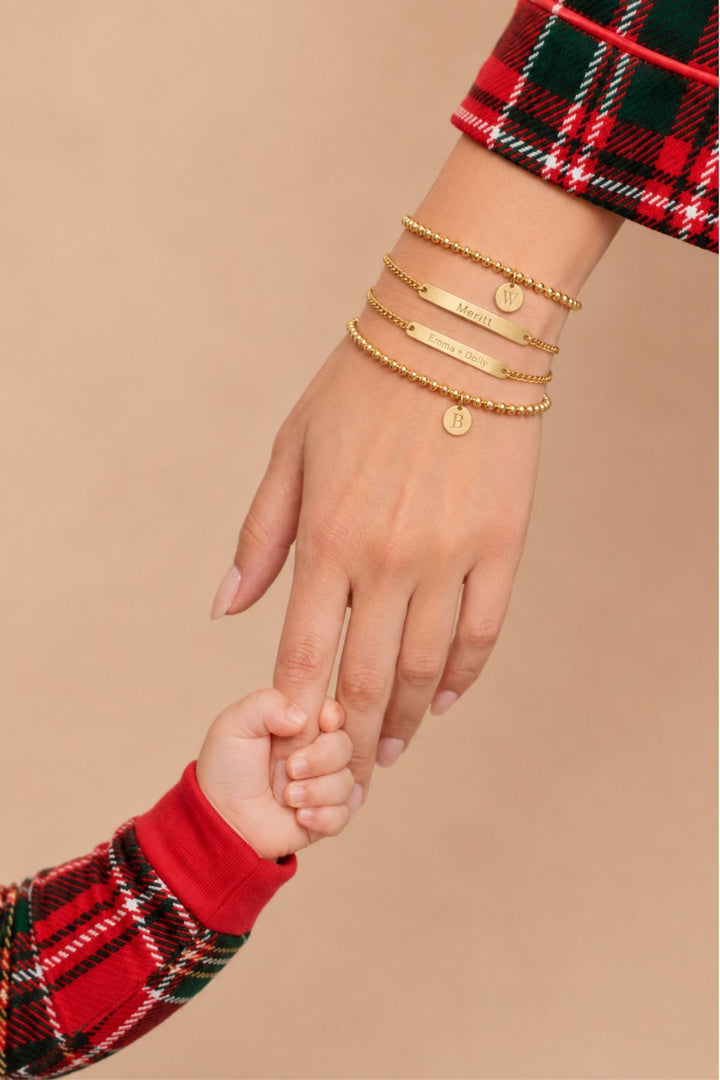 Caden Lane layered gold bracelets with "Meritt" and "Emma + Dolly" engraved bars and initial "W" and "B" discs, styled with plaid holiday pajamas, mother and child holding hands