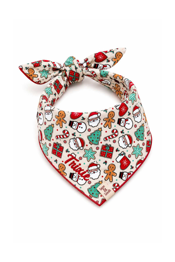Tied back Lane & Leash personalized dog bandana in cream Christmas Treats print with gingerbread men, Santa faces, snowmen, mini trees, candy canes, and gift boxes, tied bandana-style, embroidered with the name 'Trixie' in red
