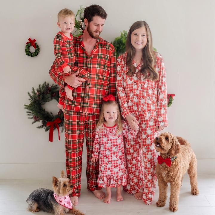 A family of four wearing Caden Lane matching red and white holiday pajamas stands with two dogs in front of a white wall decorated with green wreaths.