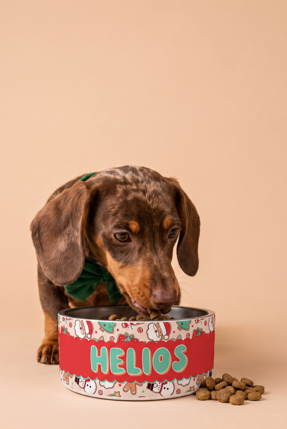 A dachshund wearing a green Lane & Leash bow tie eats from a Lane & Leash decorative red and white dog bowl with the personalized name HELIOS, set against a solid beige background with kibble scattered nearby.