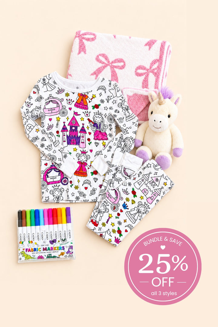 Flat lay of a Caden Lane color-in princess pajama set beside a folded pink bow blanket, paired with a plush unicorn and fabric markers.