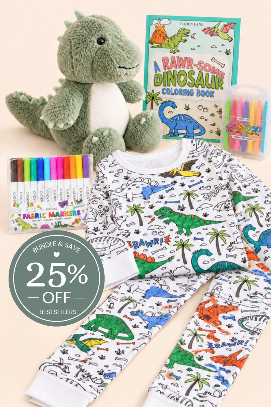 Flat lay of a Caden Lane color-in dinosaur pajama set partly filled in with marker, paired with a plush dinosaur, fabric markers, crayons, and a dinosaur coloring book.