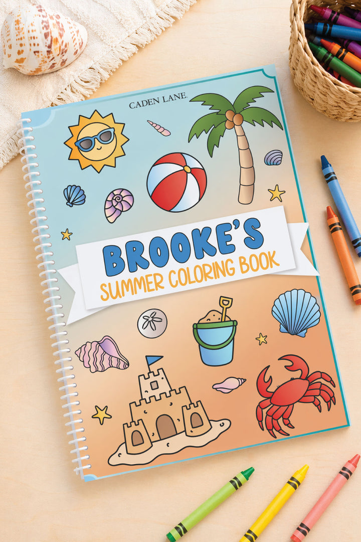 Caden Lane personalized 'Brooke's Summer Coloring Book' with beach-themed cover art, styled with a seashell, crayon basket, and scattered crayons.