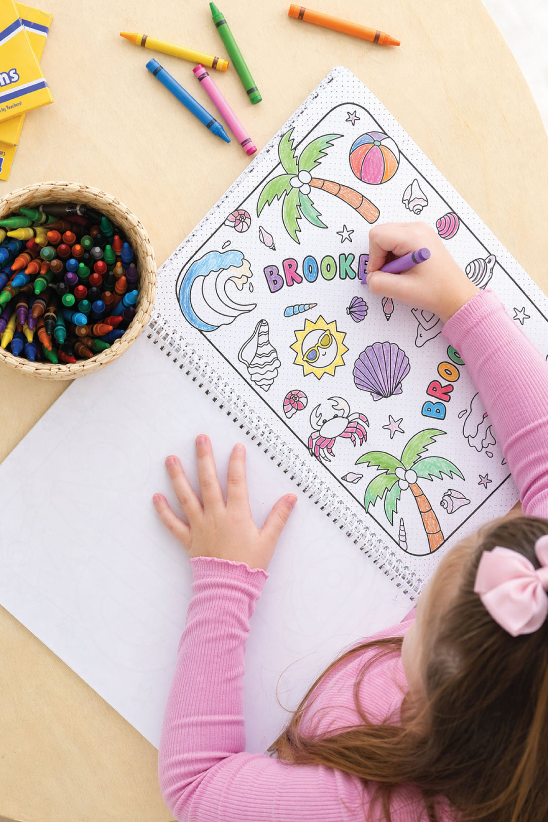 Child's hands coloring the personalized name page of a Caden Lane Summer Coloring Book with a purple marker, surrounded by crayons and a basket of crayons