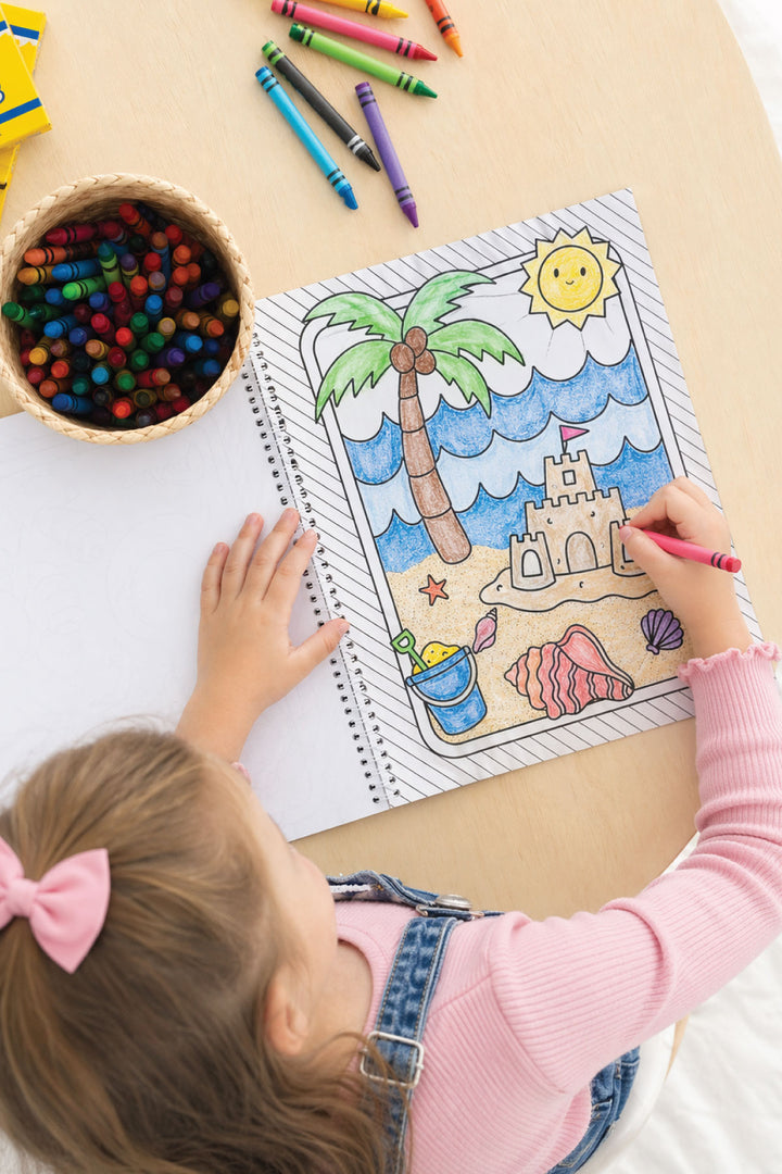 Child coloring a beach and sandcastle scene in a Caden Lane Summer Coloring Book with a pink crayon, crayon basket nearby