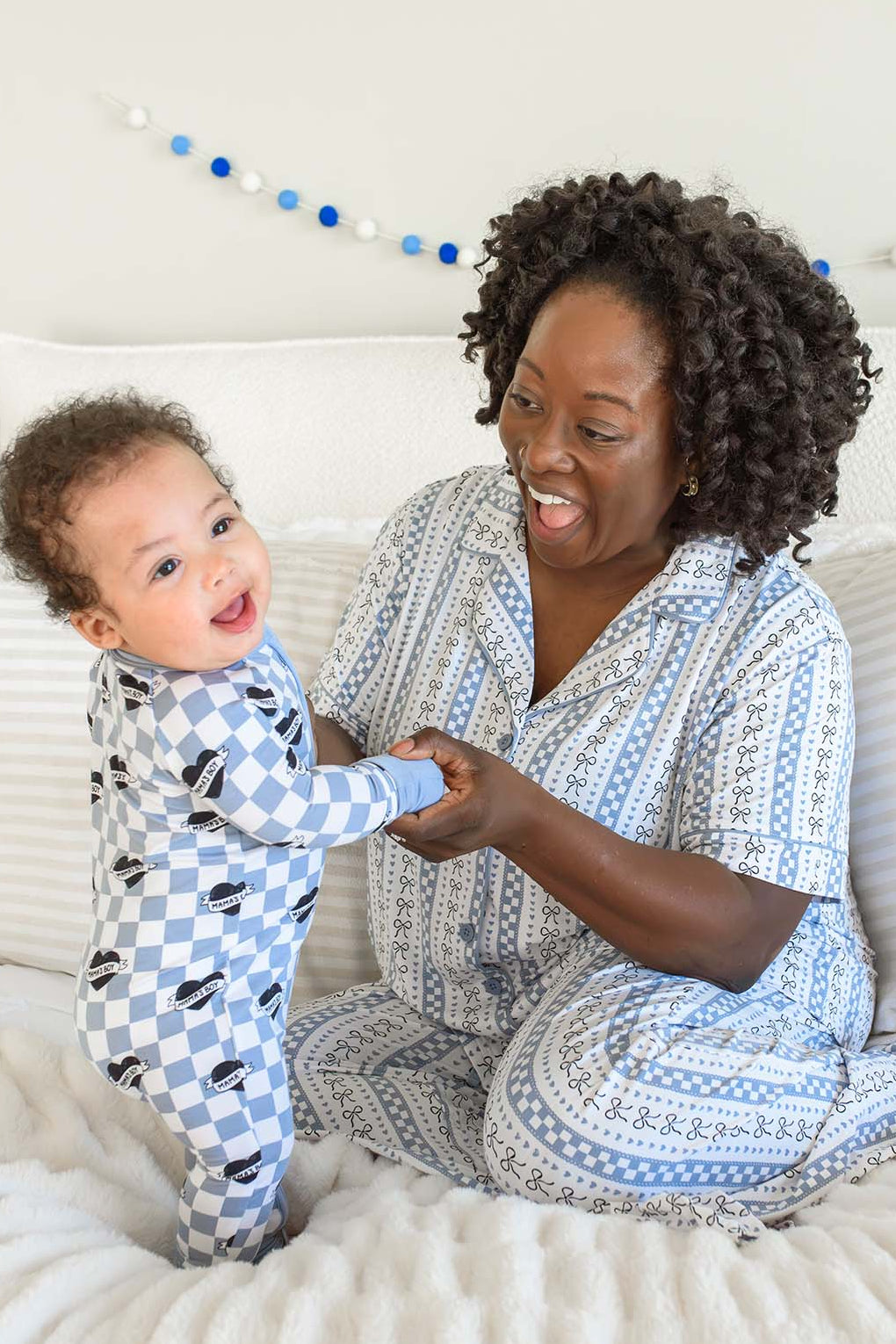 A smiling woman and a baby sitting on a bed, both wearing matching blue and white patterned pajamas.