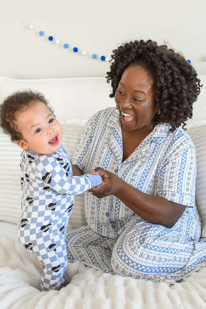 A smiling woman and a baby sitting on a bed, both wearing matching blue and white patterned pajamas.