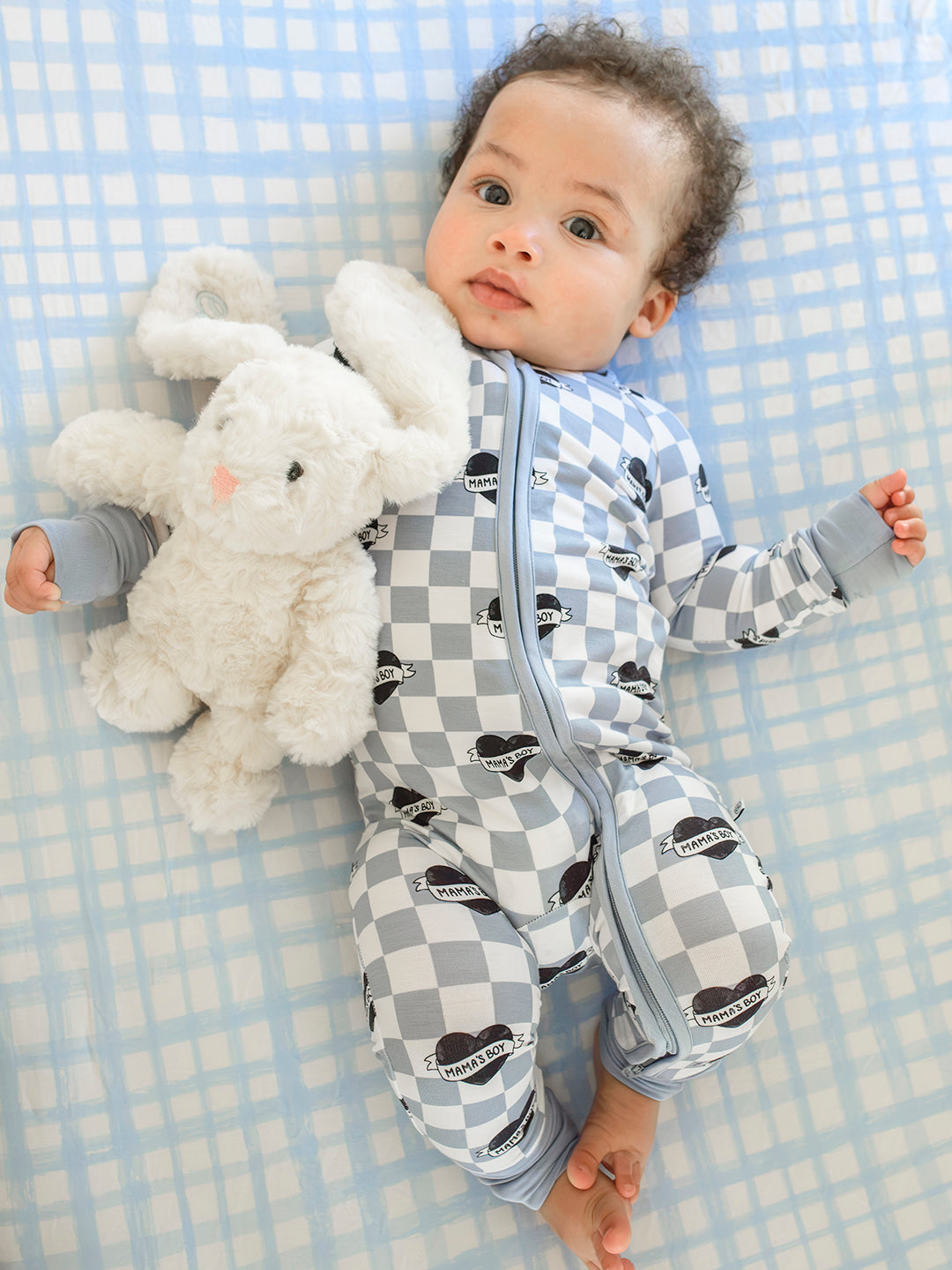 A baby lying on a blue and white checkered blanket, wearing a gray and white checkered zip-up sleeper with black heart prints, while holding a white plush bunny toy.