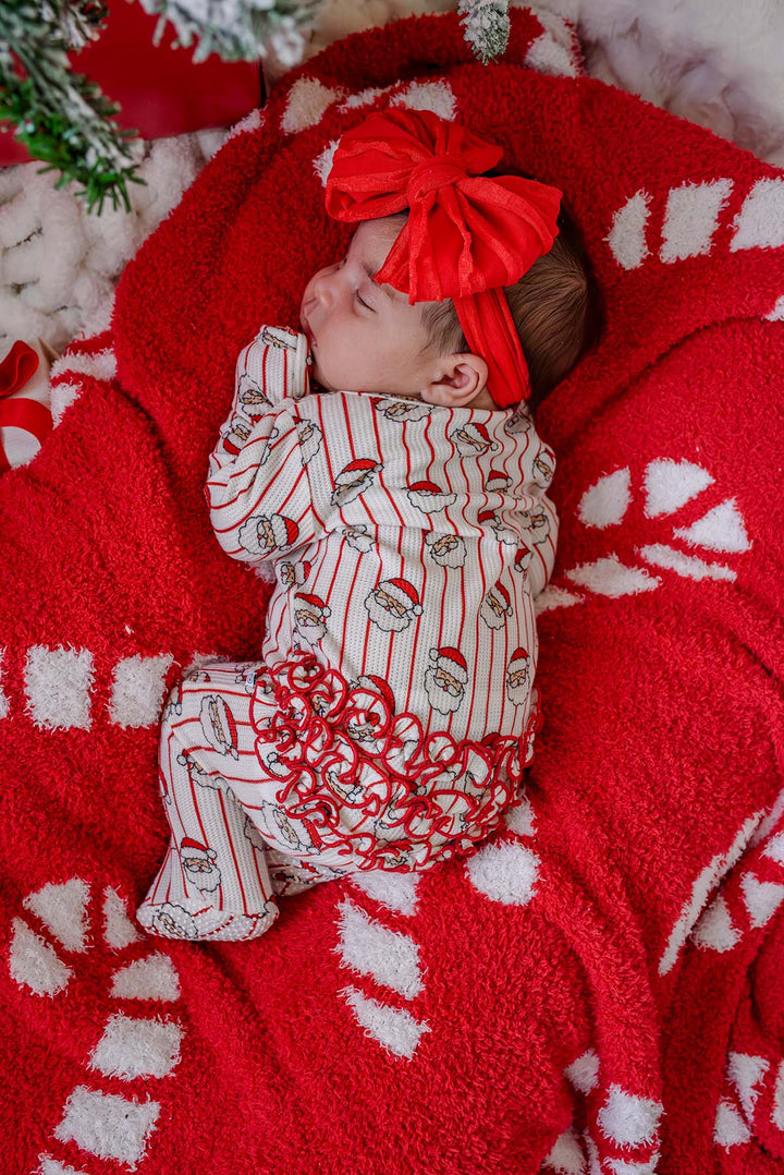 Sleeping newborn in a Santa print footie and red bow headband, resting on a Caden Lane red and white candy cane print sherpa blanket.