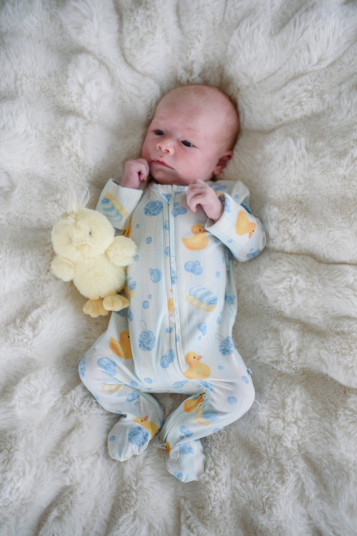 Newborn lying on white fur wearing blue Bubble Duckies bamboo zipper footie pajamas with yellow rubber duck toy