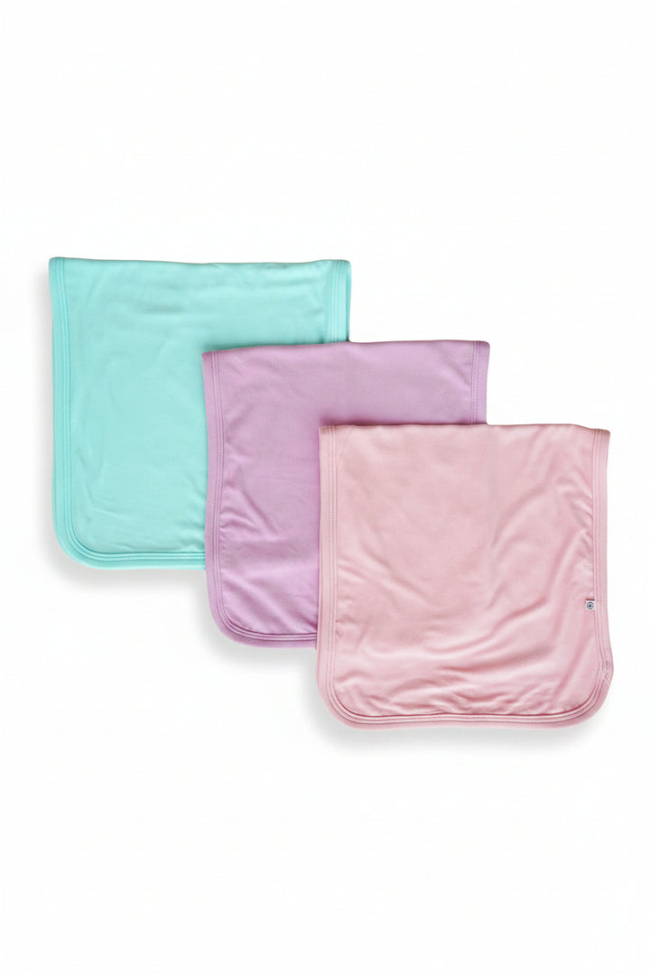 Three Caden Lane burp cloths in shades of teal, purple and pink. in pastel colors, arranged in a staggered stack on a plain white background. 