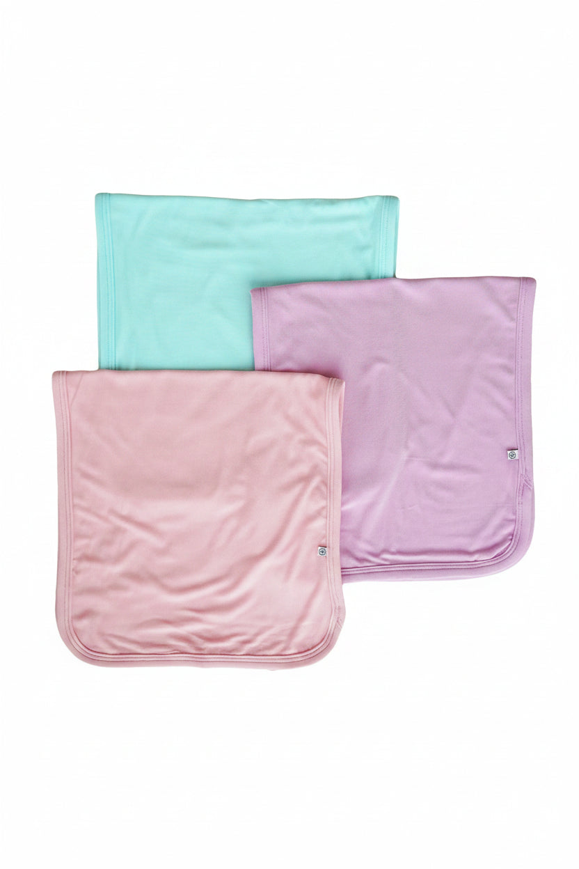 Three folded Caden Lane burp clothes in shades of teal, purple, and pink, arranged in a stack on a white background. 